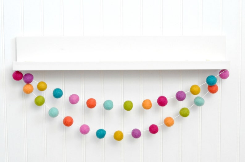 K&ouml;nnte beinhalten: Eine bunte Filzkugelgirlande mit verschiedenen Farben, darunter Rosa, Orange, Gelb, Gr&uuml;n, Blau und Lila. Die Girlande ist auf einer wei&szlig;en Schnur aufgef&auml;delt und h&auml;ngt an einem wei&szlig;en Regal.