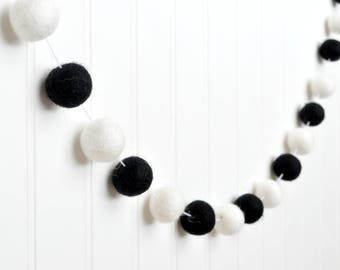 May include: A black and white felt ball garland with alternating colors. The garland is strung on a white string and is hanging on a white wall.