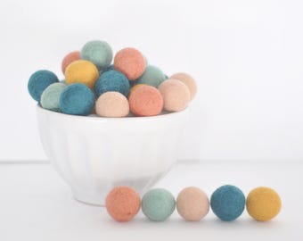 Könnte beinhalten: Eine weiße Schüssel gefüllt mit bunten Filzkugeln in den Farben Türkis, Gelb und Pfirsich. Die Schüssel steht auf einer weißen Oberfläche. Sechs Filzkugeln sind in einer Reihe auf der Oberfläche vor der Schüssel angeordnet.