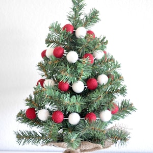 K&ouml;nnte beinhalten: Ein kleiner k&uuml;nstlicher Weihnachtsbaum, der mit einer Girlande aus roten und wei&szlig;en Filzkugeln geschm&uuml;ckt ist. Der Baum steht in einem Jutesack.