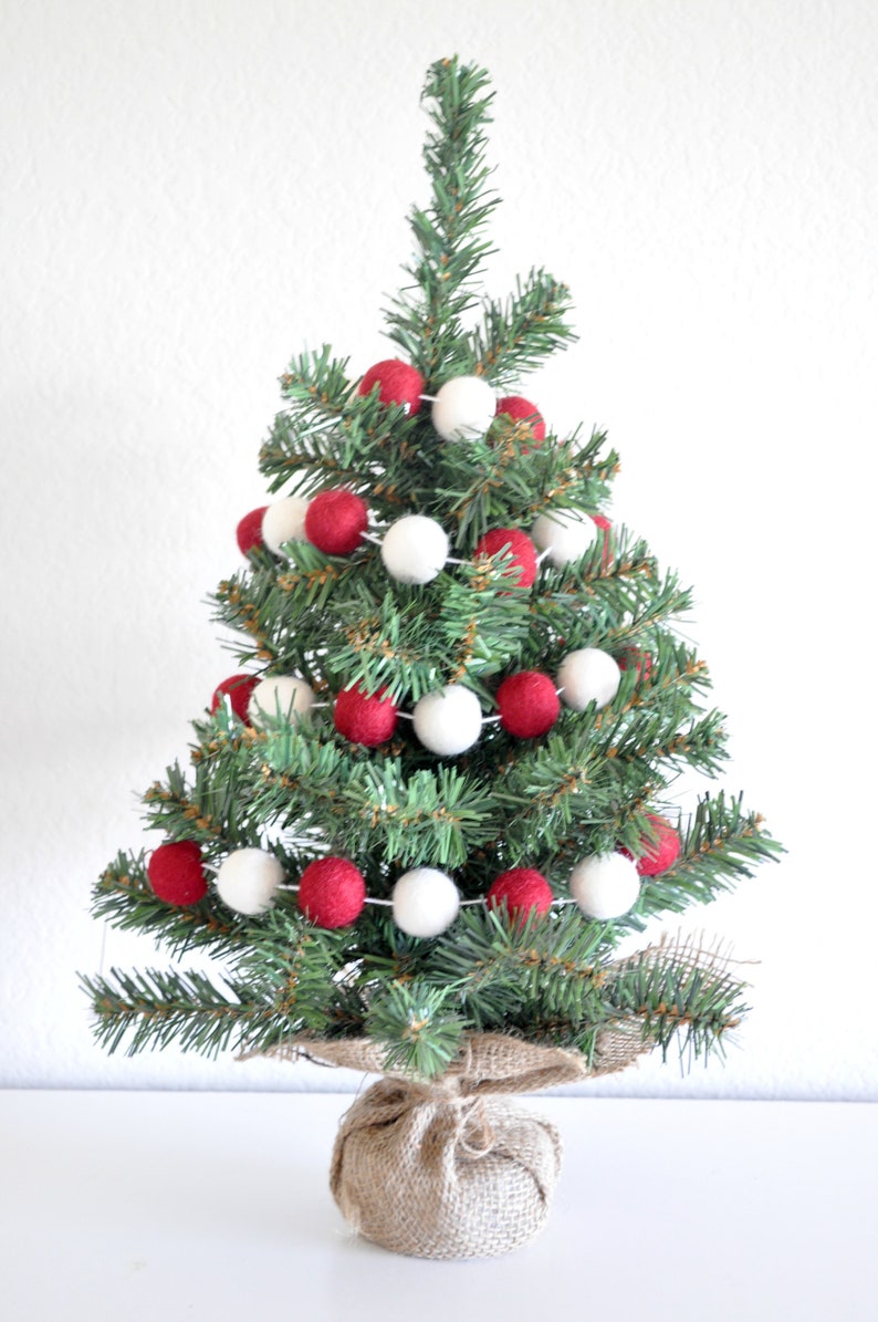 K&ouml;nnte beinhalten: Ein kleiner k&uuml;nstlicher Weihnachtsbaum, der mit einer Girlande aus roten und wei&szlig;en Filzkugeln geschm&uuml;ckt ist. Der Baum steht in einem Jutesack.