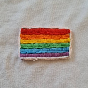 May include: Embroidered rainbow flag patch with red, orange, yellow, green, blue, and purple stripes. The patch is rectangular with a white border and is sewn onto a white fabric background. The patch is a symbol of LGBTQ+ pride.