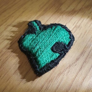 Op de afbeelding: Een groen, blad-vormig geborduurd embleem met een zwarte rand. De patch is gemaakt van textiel en ligt op een houten oppervlak. Het bladontwerp is een levendige groene kleur.