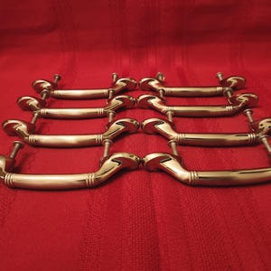 May include: Eight brass-toned drawer pulls arranged on a red background. Each pull has a curved handle with decorative end pieces and includes mounting screws. The pulls are likely for furniture restoration or new construction.
