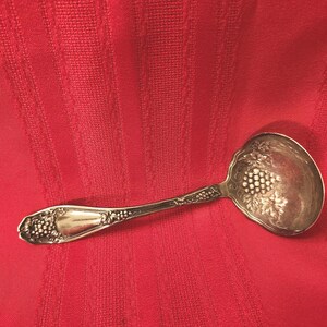 May include: Silver serving ladle with a grape design on the bowl and handle. The ladle is set against a red background. The handle has a decorative pattern. The bowl is approximately 3 inches in diameter.