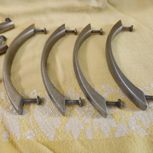 K&ouml;nnte beinhalten: Eine Sammlung antiker Schrankgriffe in Messingoptik mit gebogenen Griffen und Schrauben. Die Griffe sind auf einem gelben Stoff mit Muster angeordnet und zeigen ihr Vintage-Design und die Hardware-Details.