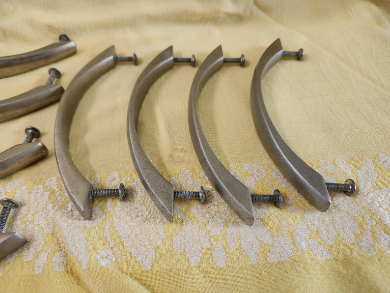 K&ouml;nnte beinhalten: Eine Sammlung antiker Schrankgriffe in Messingoptik mit gebogenen Griffen und Schrauben. Die Griffe sind auf einem gelben Stoff mit Muster angeordnet und zeigen ihr Vintage-Design und die Hardware-Details.