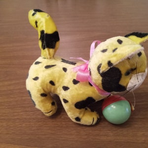 May include: A vintage plush toy cat in yellow with black spots, a pink bow, and a green and red ball. The cat has a yellow tail with black markings and black spots on its body.