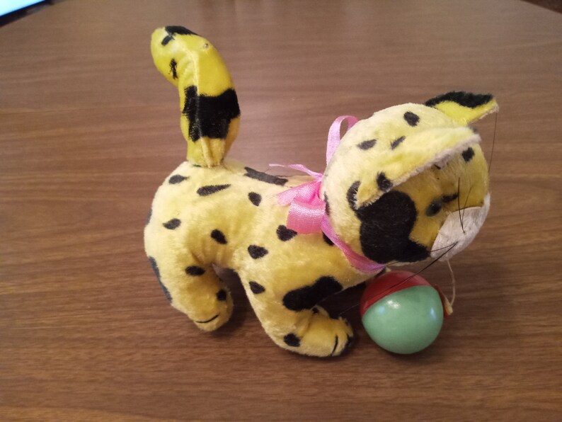 May include: A vintage plush toy cat in yellow with black spots, a pink bow, and a green and red ball. The cat has a yellow tail with black markings and black spots on its body.