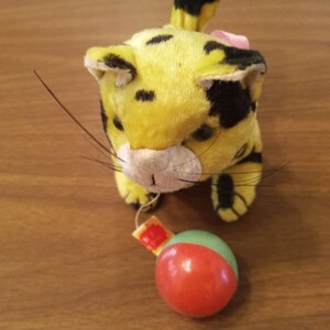 May include: A vintage plush toy cat with yellow fur and black spots, a pink bow, and long whiskers. It is paired with a red and green ball-shaped ornament, likely a Christmas decoration. The cat is positioned on a wooden surface.