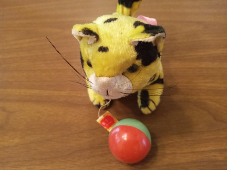 May include: A vintage plush toy cat with yellow fur and black spots, a pink bow, and long whiskers. It is paired with a red and green ball-shaped ornament, likely a Christmas decoration. The cat is positioned on a wooden surface.