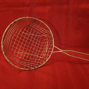 May include: A round, metal wire mesh skimmer with a long handle, ideal for straining or frying. The skimmer is made of a light-colored metal and is set against a red background.