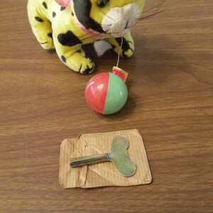 May include: A yellow plush toy leopard with black spots and a pink bow, connected to a red and green ball. A metal wind-up key rests on a piece of cardboard, suggesting a vintage toy.