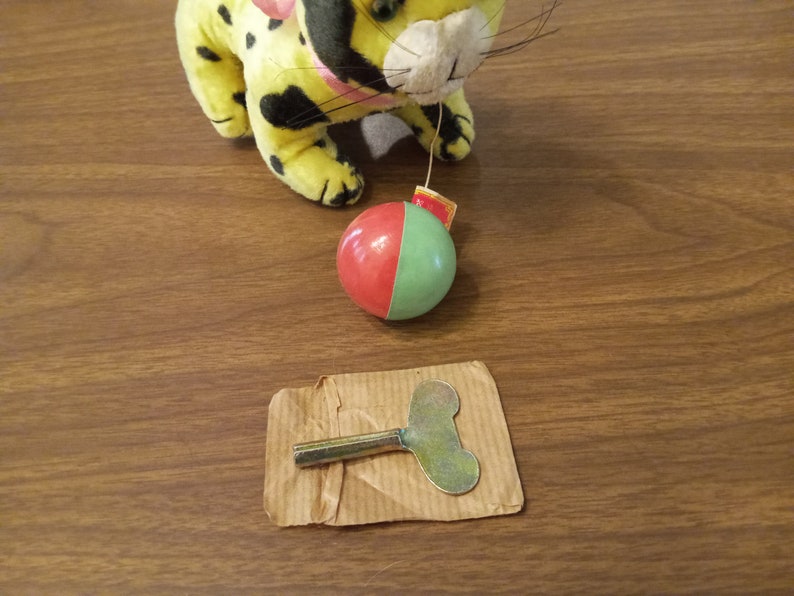 May include: A yellow plush toy leopard with black spots and a pink bow, connected to a red and green ball. A metal wind-up key rests on a piece of cardboard, suggesting a vintage toy.