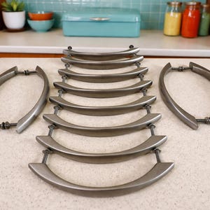 May include: Set of ten brushed nickel cabinet pulls with a curved design. The hardware is arranged on a light-colored countertop, with two pulls on the sides and eight in the center. The pulls are designed for easy installation and add a modern touch to any kitchen or bathroom.