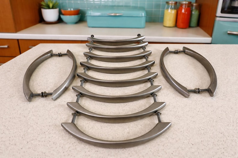 K&ouml;nnte beinhalten: Set aus zehn Schrankgriffen aus geb&uuml;rstetem Nickel mit geschwungenem Design. Die Beschl&auml;ge sind auf einer hellen Arbeitsplatte angeordnet, mit zwei Griffen an den Seiten und acht in der Mitte. Die Griffe sind f&uuml;r eine einfache Montage konzipiert.