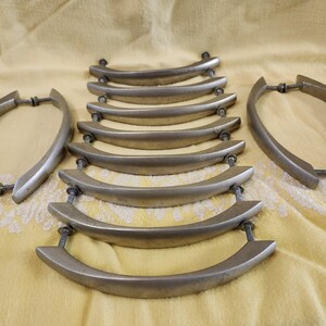 K&ouml;nnte beinhalten: Eine Sammlung silberfarbener Schrankgriffe mit gebogenem Design. Die Griffe sind auf einer gelben Stoffoberfl&auml;che angeordnet. Jeder Griff hat zwei Schraubbefestigungen, f&uuml;r einen Vintage-Look.