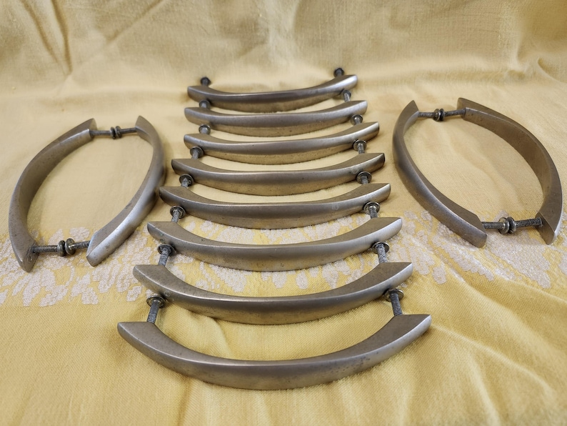 K&ouml;nnte beinhalten: Eine Sammlung silberfarbener Schrankgriffe mit gebogenem Design. Die Griffe sind auf einer gelben Stoffoberfl&auml;che angeordnet. Jeder Griff hat zwei Schraubbefestigungen, f&uuml;r einen Vintage-Look.