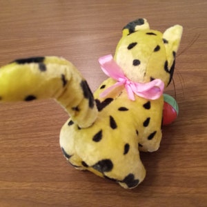 May include: A vintage plush toy cat in yellow with black spots, a pink bow, and a small ball. The cat's tail is curved, and it has a playful appearance. The toy is likely from the mid-20th century.