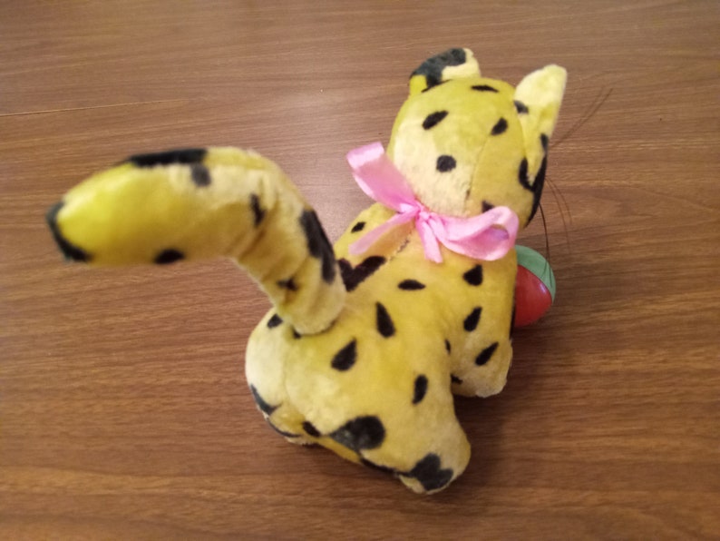 May include: A vintage plush toy cat in yellow with black spots, a pink bow, and a small ball. The cat's tail is curved, and it has a playful appearance. The toy is likely from the mid-20th century.