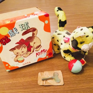 May include: Vintage wind-up toy cat with a yellow and black spotted body, a red and white ball, and a box with a cat illustration. The box is orange with Chinese characters and the word "Kitty".