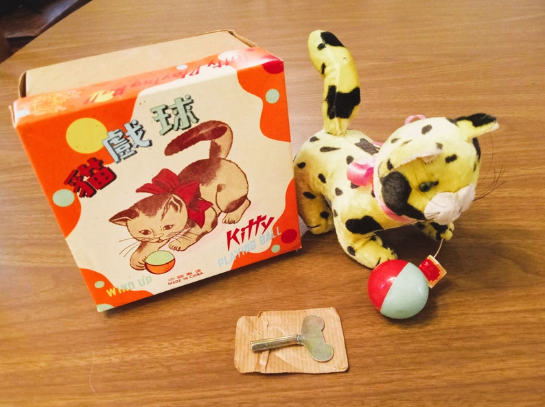 May include: Vintage wind-up toy cat with a yellow and black spotted body, a red and white ball, and a box with a cat illustration. The box is orange with Chinese characters and the word "Kitty".