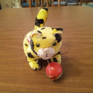May include: A plush toy cat in yellow with black spots, playing with a red ball. The cat has a white muzzle, black whiskers, and a striped tail. The toy is on a wooden surface.