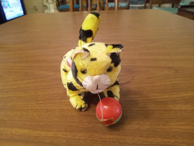 May include: A plush toy cat in yellow with black spots, playing with a red ball. The cat has a white muzzle, black whiskers, and a striped tail. The toy is on a wooden surface.