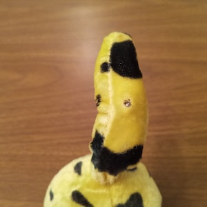 May include: A plush toy with a yellow body and black spots. The toy has a curved shape with a small, rounded head. The toy has two small, shiny embellishments on its head.
