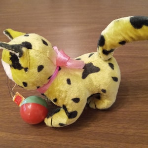 May include: A vintage plush toy cat in yellow with black spots, a pink ribbon, and a small red and green ball. The cat has a long tail and is posed on a wooden surface. The toy is a classic children's item.