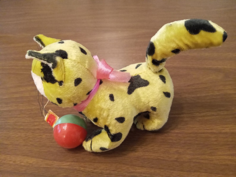 May include: A vintage plush toy cat in yellow with black spots, a pink ribbon, and a small red and green ball. The cat has a long tail and is posed on a wooden surface. The toy is a classic children's item.