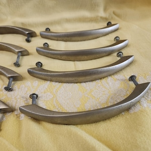 K&ouml;nnte beinhalten: Eine Sammlung von Schrankgriffen im antiken Stil. Diese gebogenen Metallgriffe haben eine geb&uuml;rstete Bronzeoberfl&auml;che und sind mit Schrauben befestigt. Die Hardware ist auf einer gelben Oberfl&auml;che mit floralem Muster angeordnet.