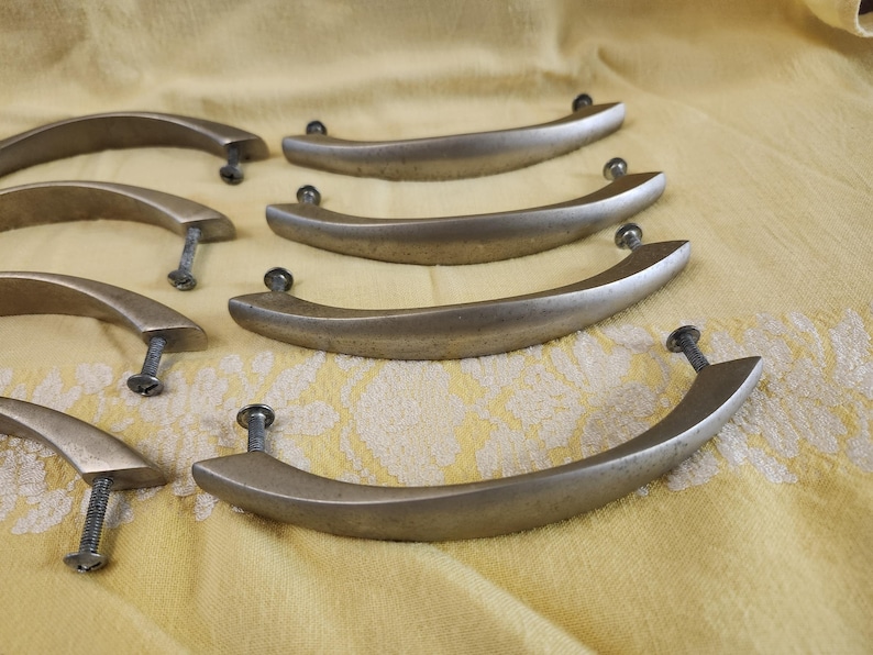 K&ouml;nnte beinhalten: Eine Sammlung von Schrankgriffen im antiken Stil. Diese gebogenen Metallgriffe haben eine geb&uuml;rstete Bronzeoberfl&auml;che und sind mit Schrauben befestigt. Die Hardware ist auf einer gelben Oberfl&auml;che mit floralem Muster angeordnet.