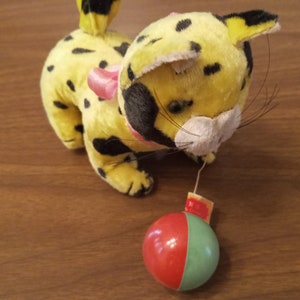 May include: A vintage plush toy leopard with yellow fur and black spots. It has a pink bow and a red and green ball attached by a string. The toy is on a wooden surface.