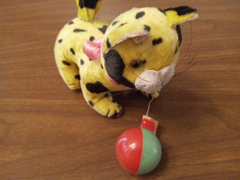 May include: A vintage plush toy leopard with yellow fur and black spots. It has a pink bow and a red and green ball attached by a string. The toy is on a wooden surface.