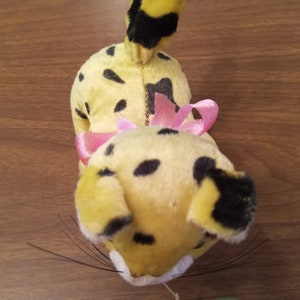 May include: A vintage plush toy cheetah with yellow fur, black spots, and a pink bow. The cheetah has black whiskers and a black-tipped tail. The toy is positioned on a wooden surface.