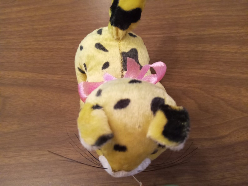 May include: A vintage plush toy cheetah with yellow fur, black spots, and a pink bow. The cheetah has black whiskers and a black-tipped tail. The toy is positioned on a wooden surface.