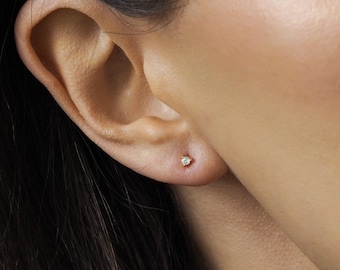 Puede incluir: Un pequeño pendiente de oro con una sola gema transparente se muestra en un piercing en la oreja. El pendiente es redondo y está engastado en un engaste de oro. La piel alrededor de la oreja es visible.