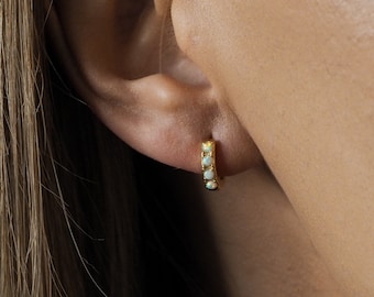 May include: Close-up of a gold hoop earring with five small, round, opalescent stones. The earring is worn on an ear, with the skin tone in the background. The earring has a simple, elegant design.