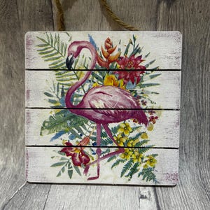 May include: A square wooden wall sign featuring a printed illustration of a pink flamingo surrounded by vibrant tropical foliage and flowers, including red and yellow blossoms, with a thin white heart outline on the flamingo's body.