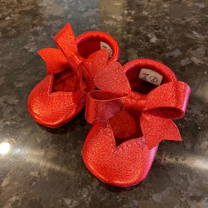 K&ouml;nnte beinhalten: Ein Paar rote Lederschuhe f&uuml;r Babys mit einer gro&szlig;en Schleife an jedem Schuh. Die Schuhe sind in den USA hergestellt.