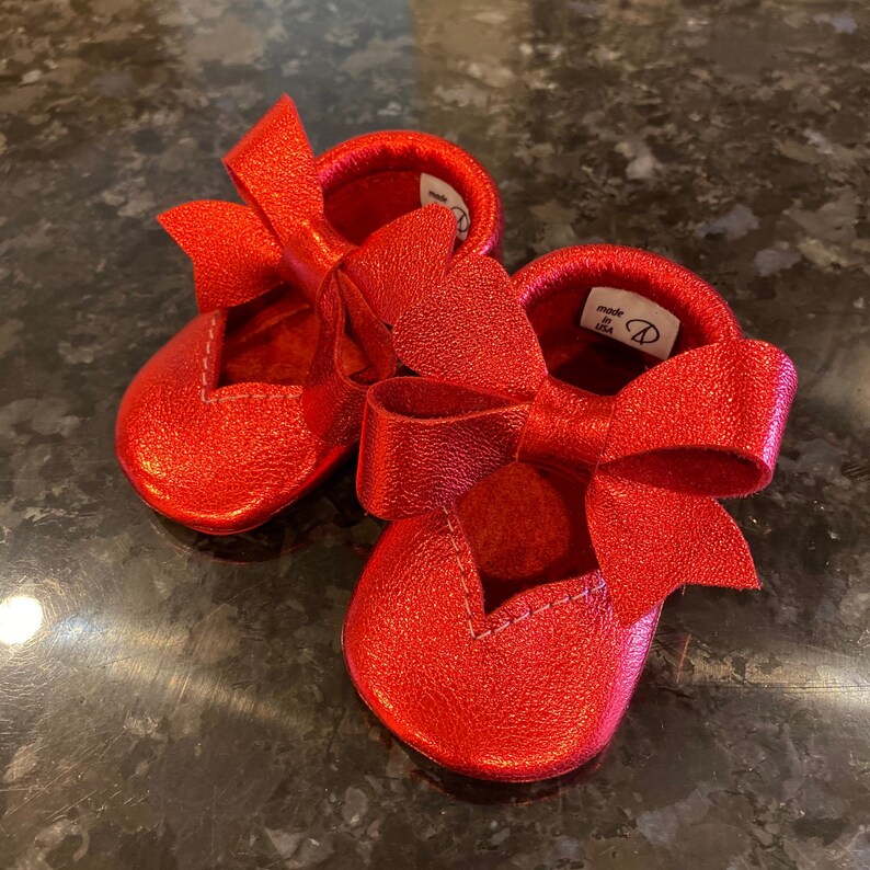 K&ouml;nnte beinhalten: Ein Paar rote Lederschuhe f&uuml;r Babys mit einer gro&szlig;en Schleife an jedem Schuh. Die Schuhe sind in den USA hergestellt.