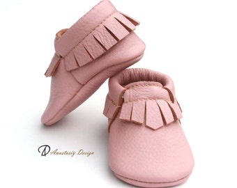 May include: A pair of pink leather baby moccasins with fringe detailing. The moccasins are soft and flexible, perfect for little feet.