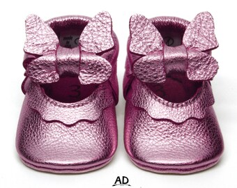 May include: Pink leather baby shoes with a butterfly bow detail. The shoes have a scalloped edge and a metallic finish.