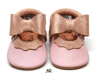 May include: Pink leather baby shoes with a gold glitter bow. The shoes have a scalloped edge and are made in the USA.