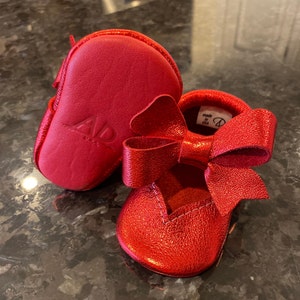 K&ouml;nnte beinhalten: Rote Lederschuhe f&uuml;r Babys mit einer Schleife auf der Vorderseite. Die Schuhe sind in den USA hergestellt und tragen den Markennamen "AID" an der Seite.