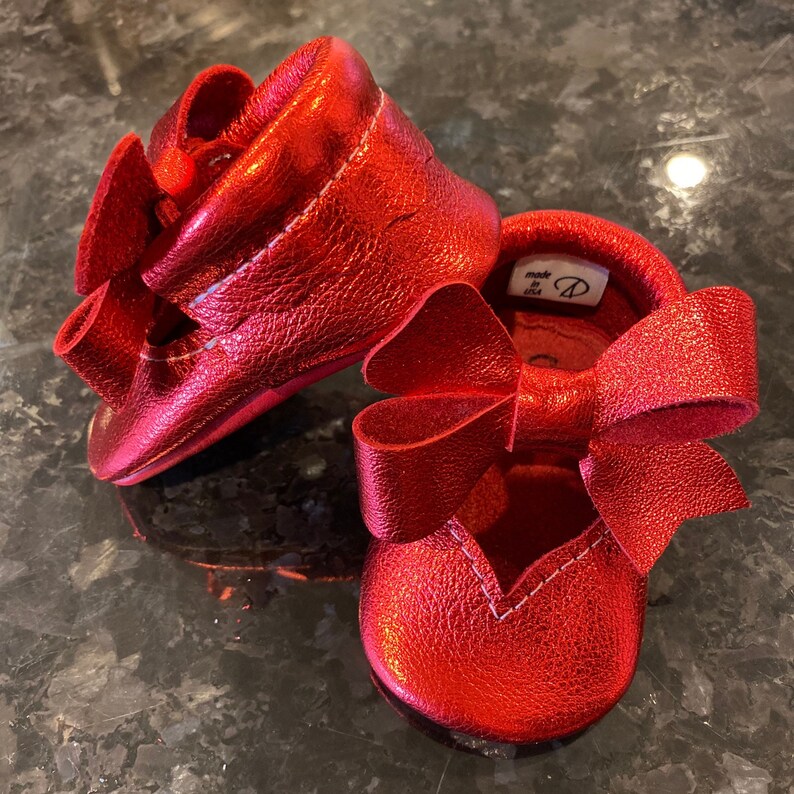 K&ouml;nnte beinhalten: Ein Paar rote Lederschuhe f&uuml;r Babys mit einer Schleife an jedem Schuh. Die Schuhe sind in den USA hergestellt.