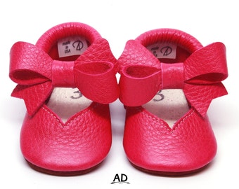 May include: A pair of bright pink leather baby shoes with a large bow on the front. The shoes are made in the USA.