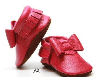 May include: A pair of pink leather baby moccasins with a bow on the front. The moccasins have a fringe detail on the back.
