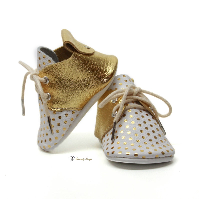 Puede incluir: Un par de zapatos de beb&eacute; de cuero blanco con lunares dorados. Un zapato es de cuero dorado con lunares blancos. Los zapatos tienen cordones y est&aacute;n atados.